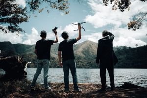 Three men holding drones by a mountain lake, silhouette against a scenic backdrop.